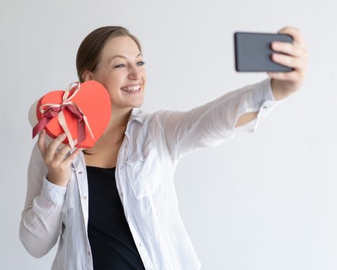 Cheerful woman taking selfie photo with heart shaped gift box. Lady wearing casual shirt and posing. Valentines Day and technology concept. Isolated view on white background.