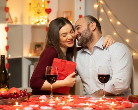 young beautiful couple man and woman with present sitting at the table decorated with candles and rose petals happy in love celebrating international women's day march in decorated living room