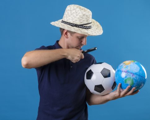 young handsome man in summer hat holding soccer ball and globe looking through magnifying glass on them intrigued standing over blue background